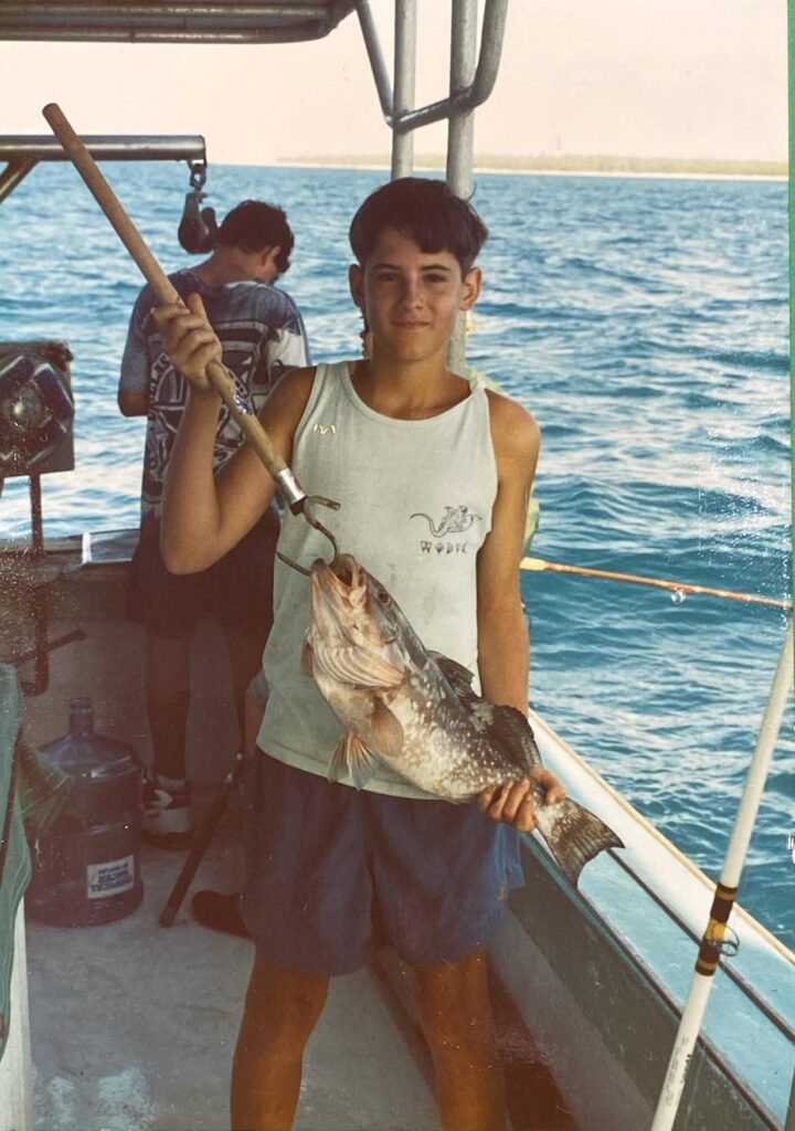 Old picture of a kid in a tank top holding a red grouper with a gaff through its mouth.