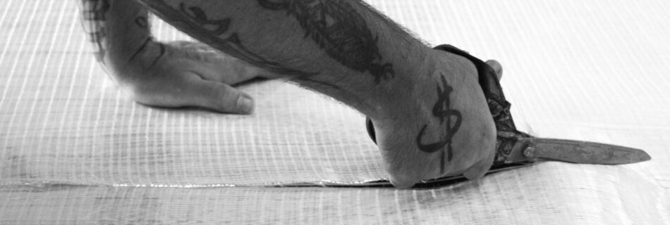 A hand with a tattoo of a dollar sign cuts through fiberglass cloth